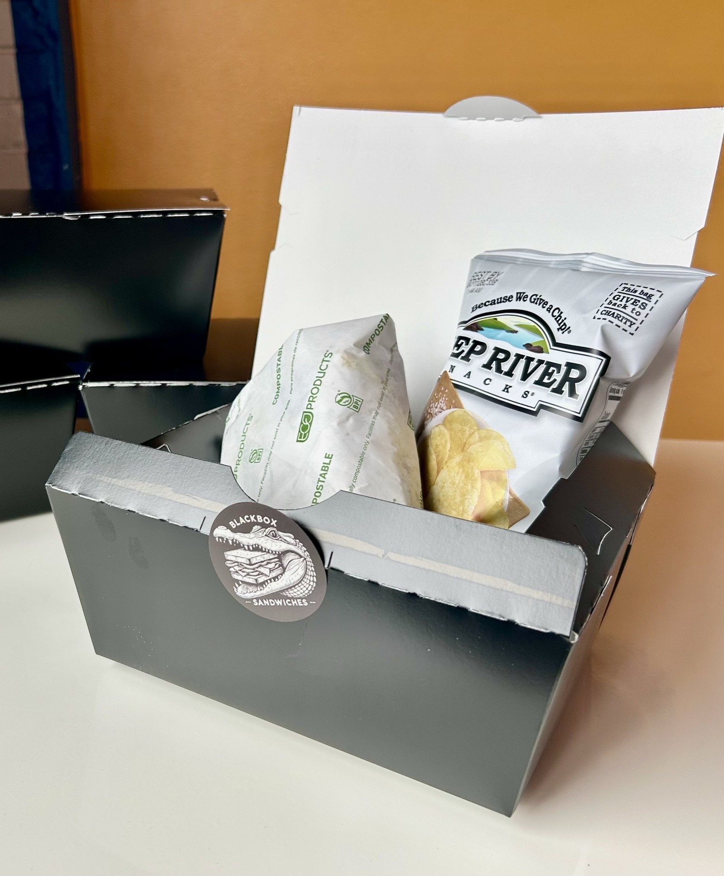A black takeout box open on a white counter: a sandwich in compostable wrap and a bag of Deep River chips inside, the logo stickered onto the front flap, and a stack of closed black boxes behind it against an orange wall.