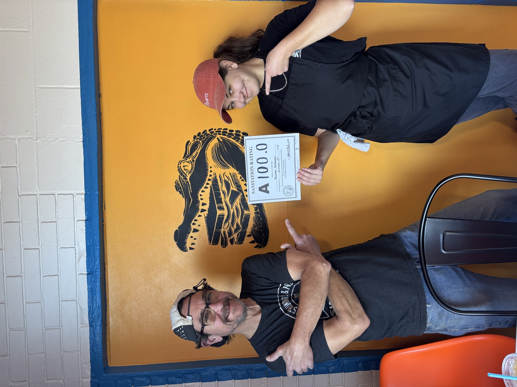 Two people standing in front of the yellow alligator panel in the dining room, arms folded, holding up a health inspection certificate scored 100.