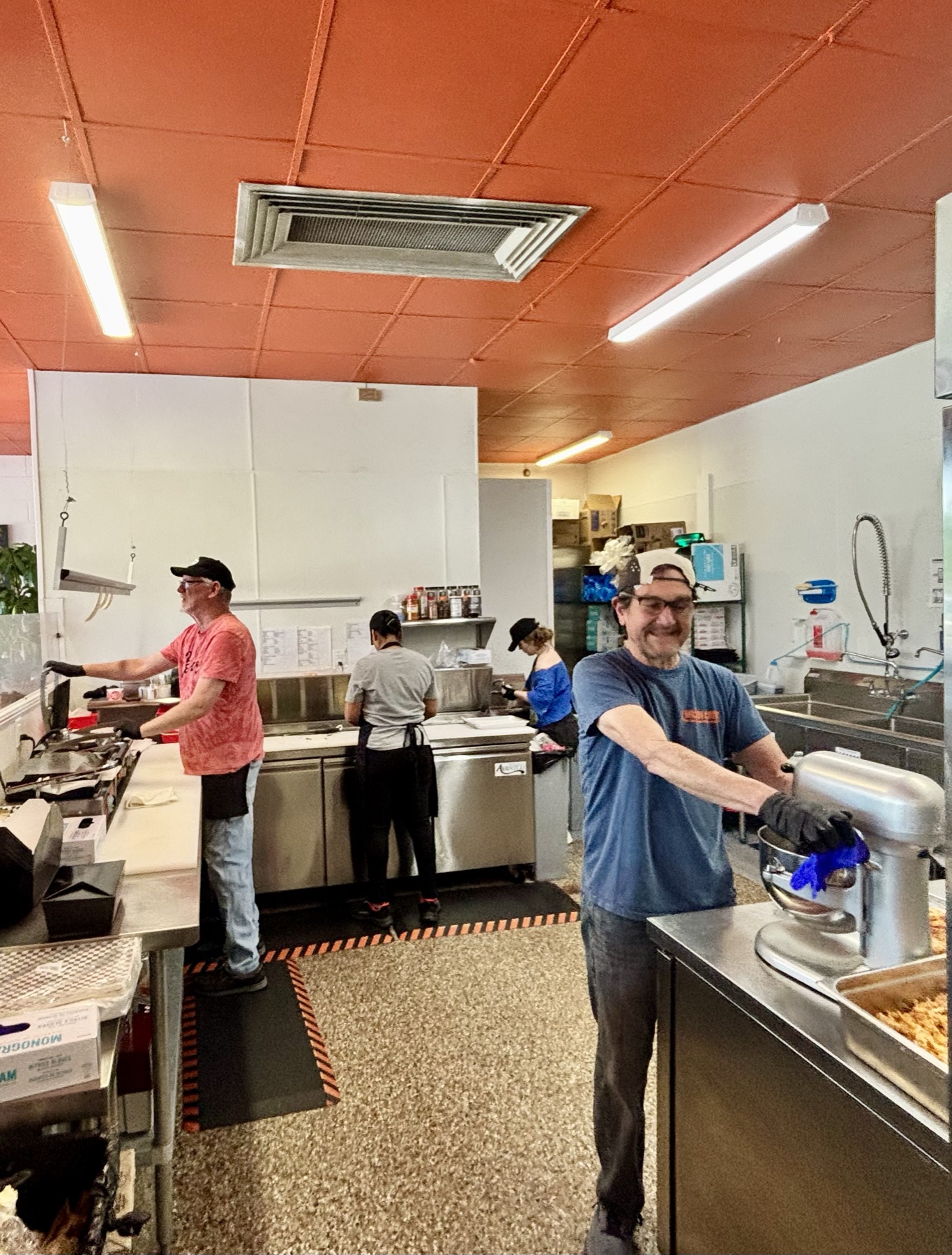 The kitchen seen from the register position, under the orange ceiling. A cook in red works the presses in the corner; behind him the builder in gray stands at the make unit; another person works at the back bench, and a fourth runs a stand mixer at the prep counter.