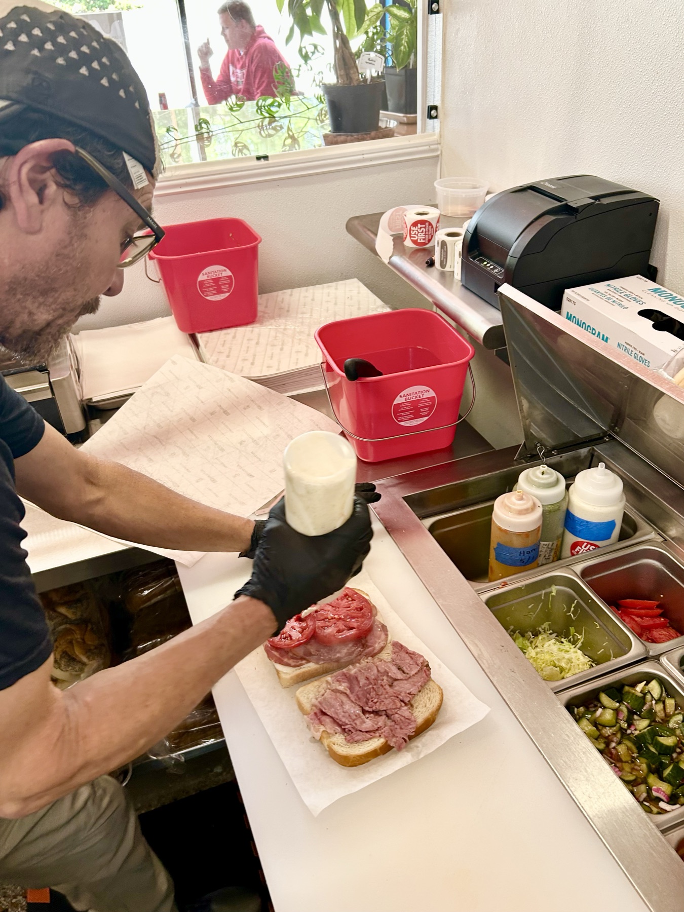 The same photograph of the line, read for its detail: two red sanitation buckets, one with a knife handle standing in it; a box of nitrile gloves on the shelf beside a receipt printer; two rolls of red USE FIRST stickers; a stack of sandwich wrap; and squeeze bottles labeled and dated in marker, one carrying a USE FIRST sticker, above pans of shredded lettuce, sliced tomato and house pickle.