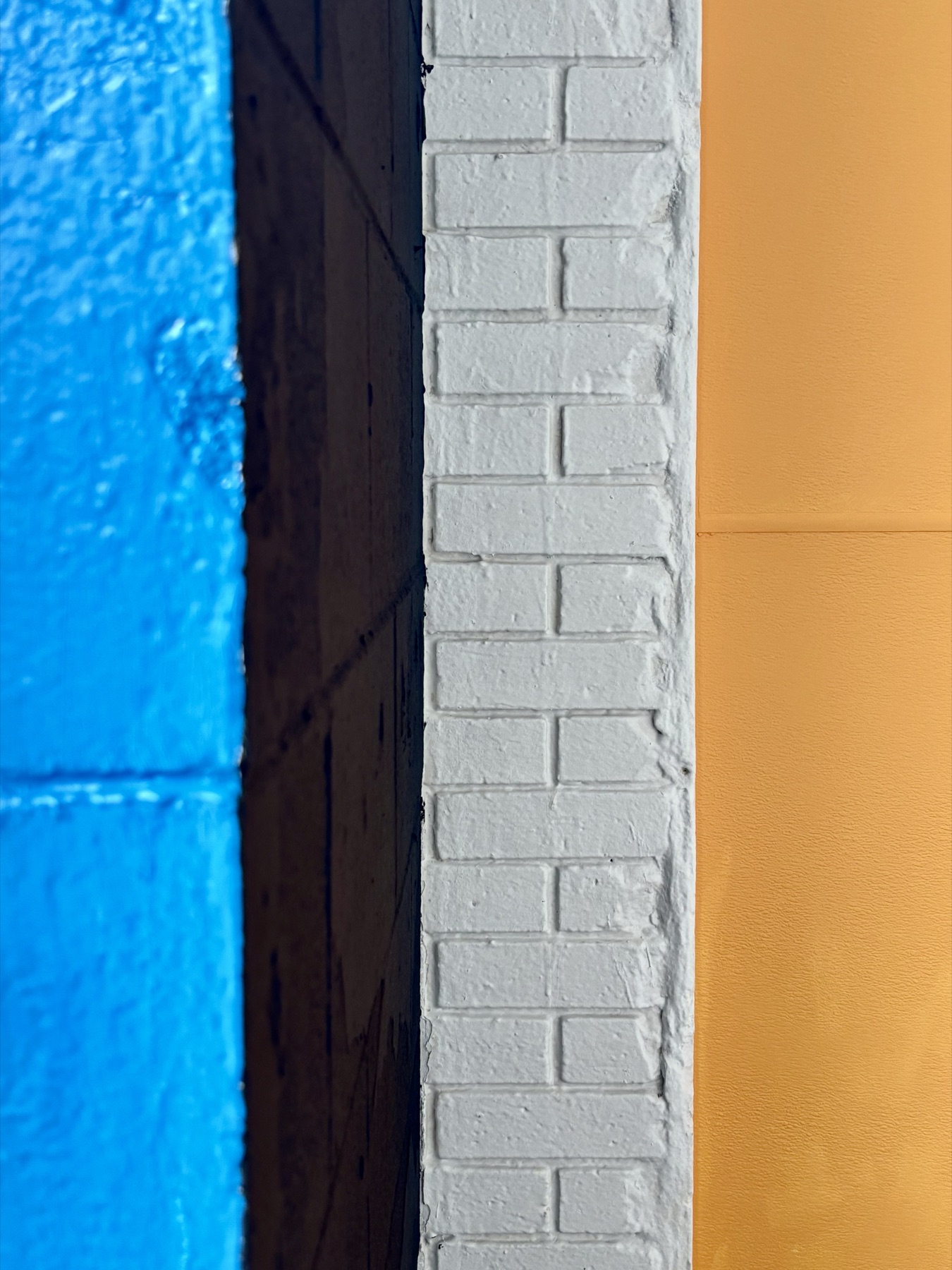 A close view of four surfaces meeting: a blue-painted block wall, a black reveal, a white-painted block pier, and a flat orange wall, in four vertical bands.