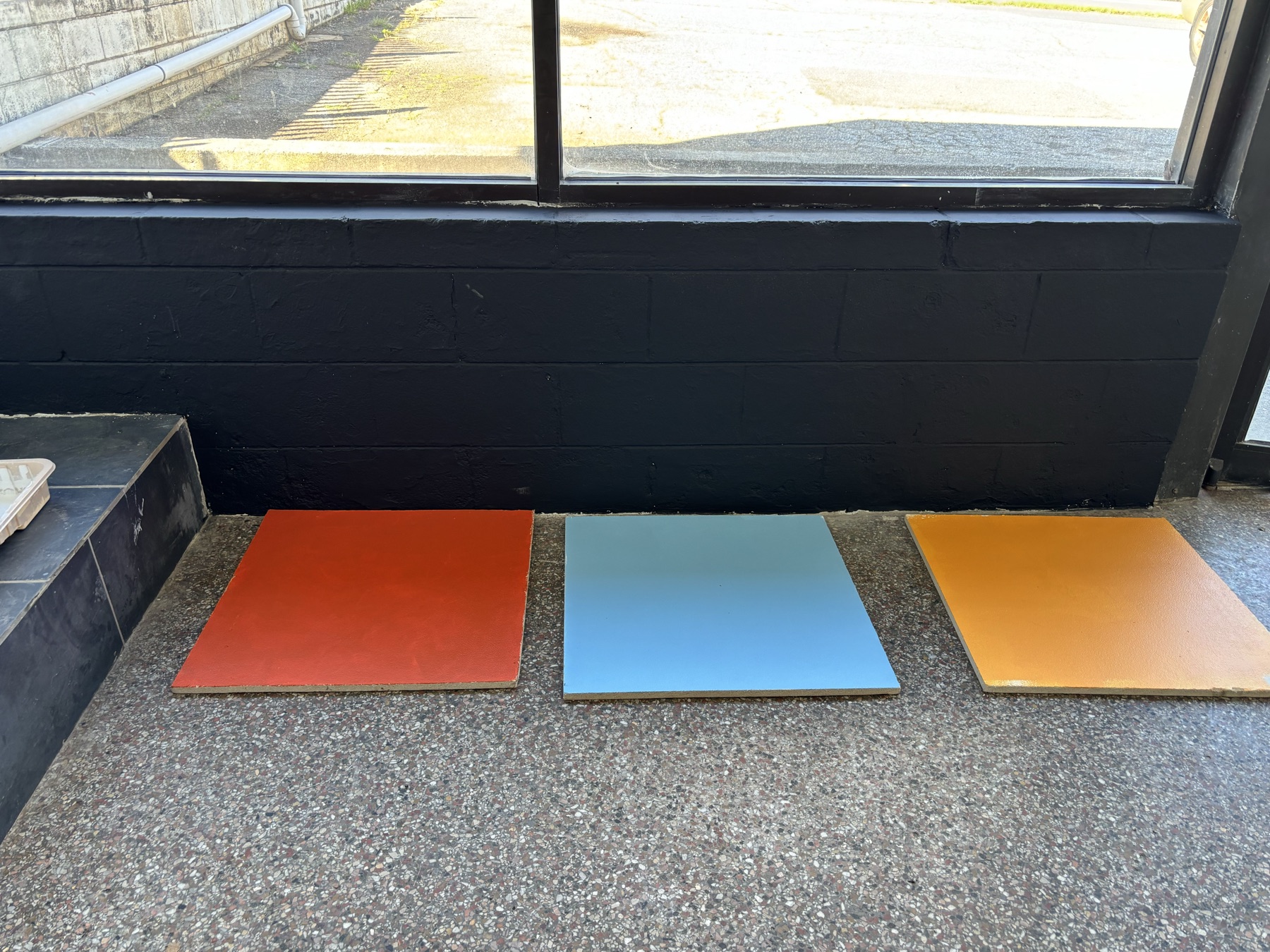 Three ceiling tiles painted as samples and laid on the existing speckled floor under the front window: a burnt orange, a mid blue and a warmer orange, against the black-painted base of the block wall.