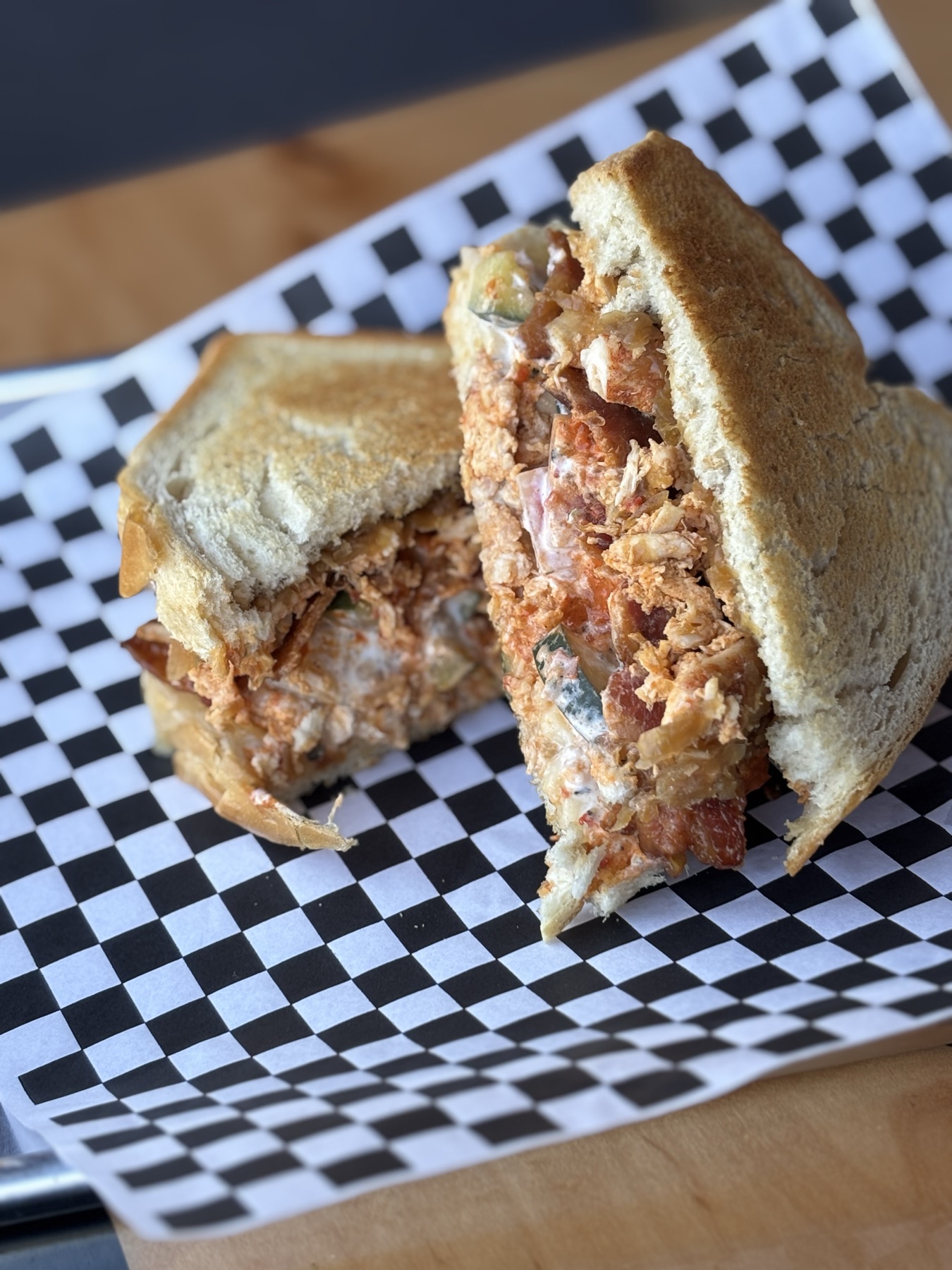A chopped sandwich on a roll, halved and standing on checkered paper on a wooden table by a window.