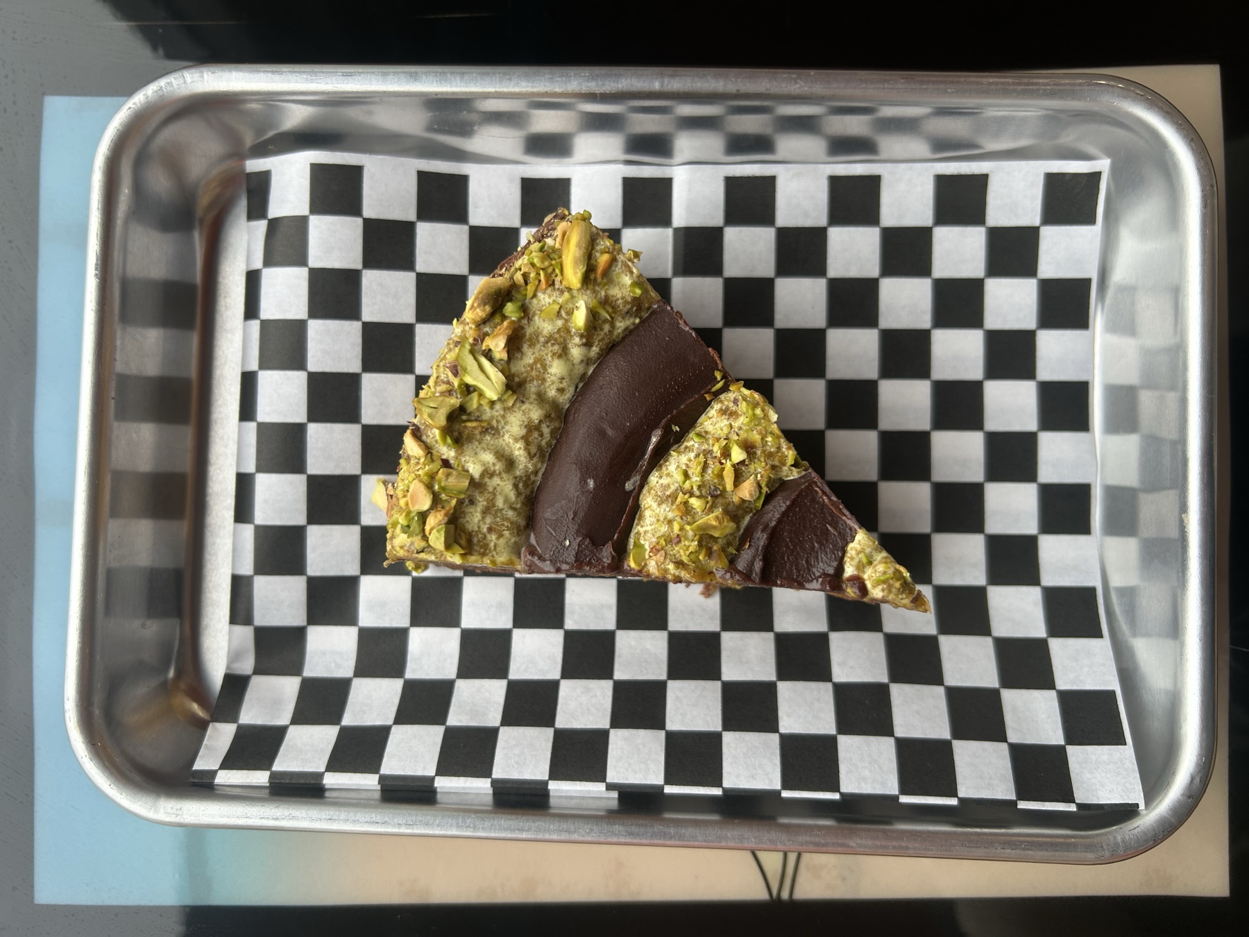 A slice of pistachio and chocolate dessert on checkered paper, on a stainless tray.