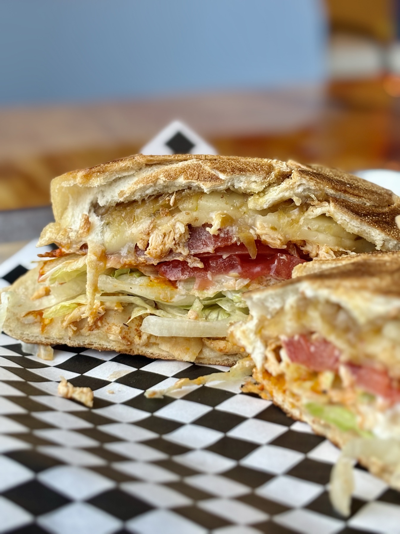 A pressed sandwich cut in half and stood on its cut edges: toasted bread, melted cheese, shredded chicken, tomato and iceberg showing at the cut, on black and white checkered paper.