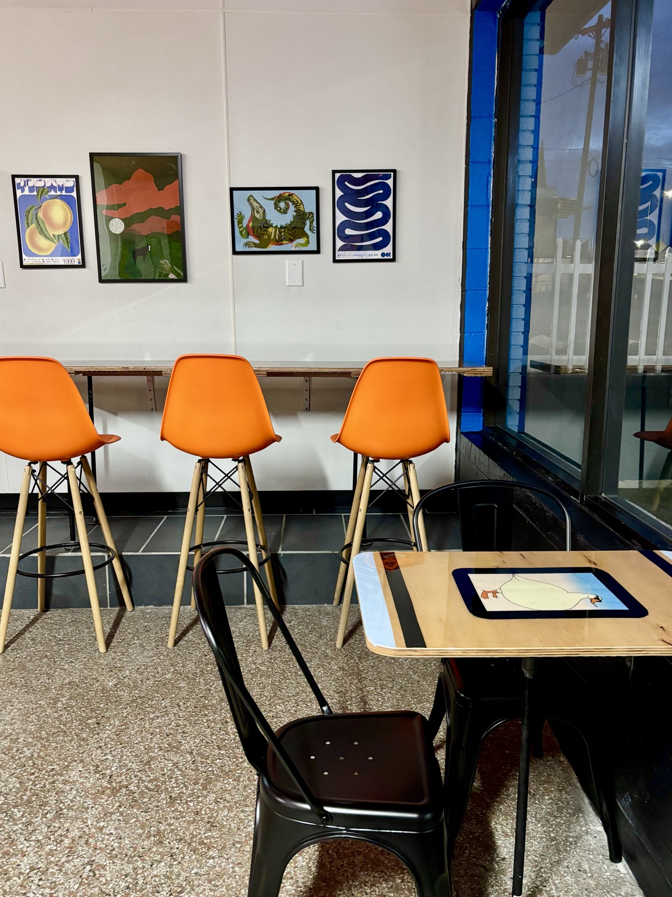 Orange molded stools along a plywood counter at the window, with four framed prints above and a small table carrying a goose illustration set under its surface.