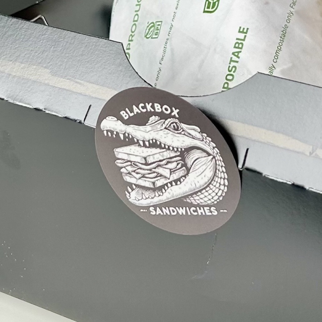 A round sticker roughly an inch and a half across, sealing the front flap of a black takeout box: the alligator and sandwich in cream on a dark ground, the fine linework still legible at that size.