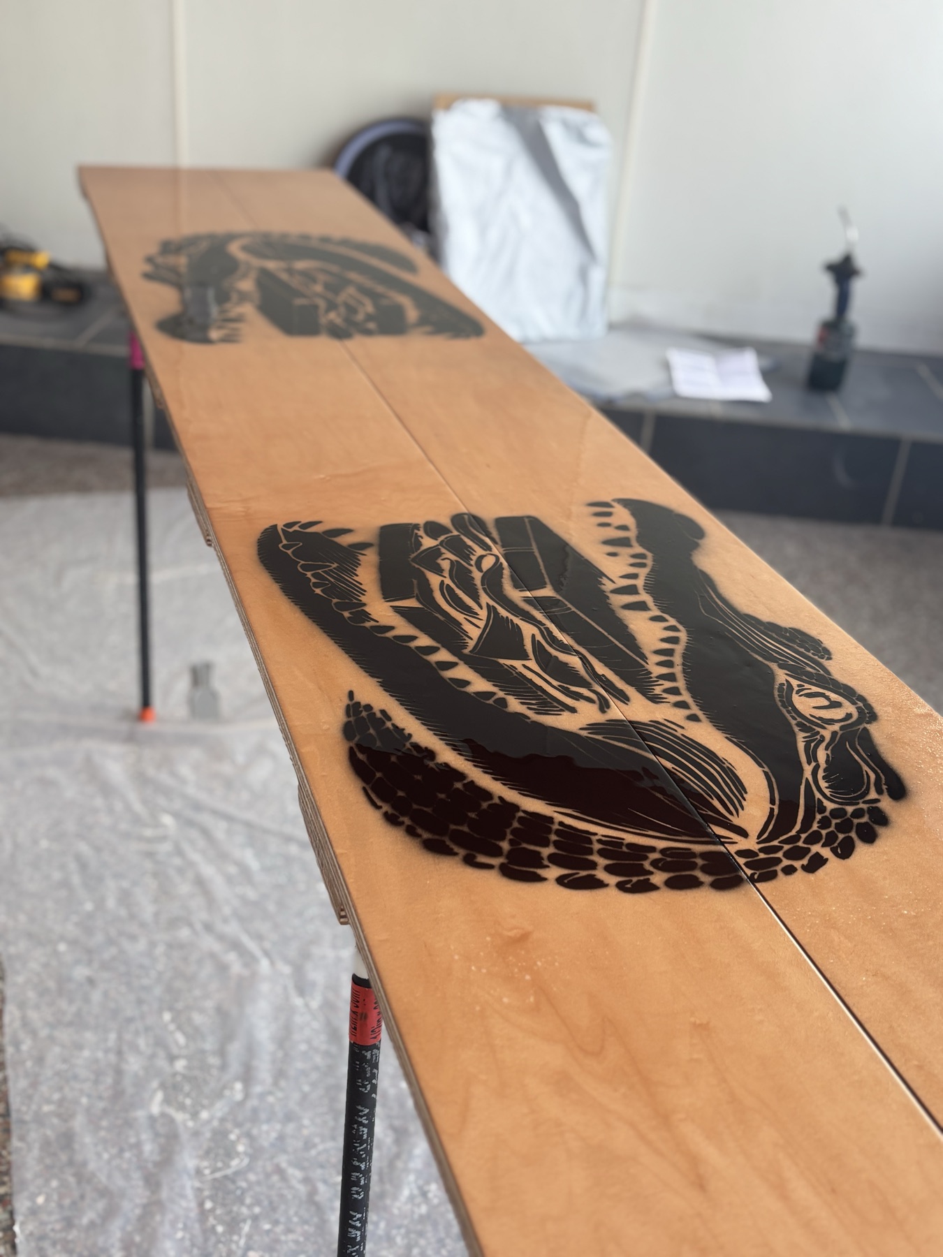 The long wall table on trestles, its plywood top finished in warm orange, with two alligator logos sprayed across it in black through a stencil, each roughly three feet long.