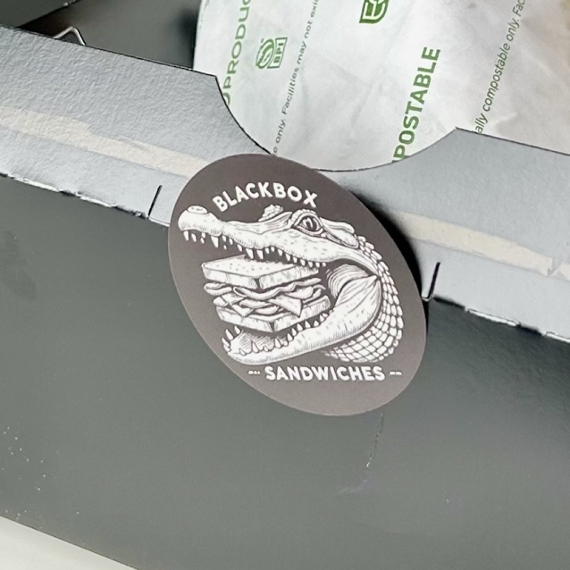 A round sticker roughly an inch and a half across, sealing the front flap of a black takeout box: the alligator and sandwich in cream on a dark ground, the fine linework still legible at that size.